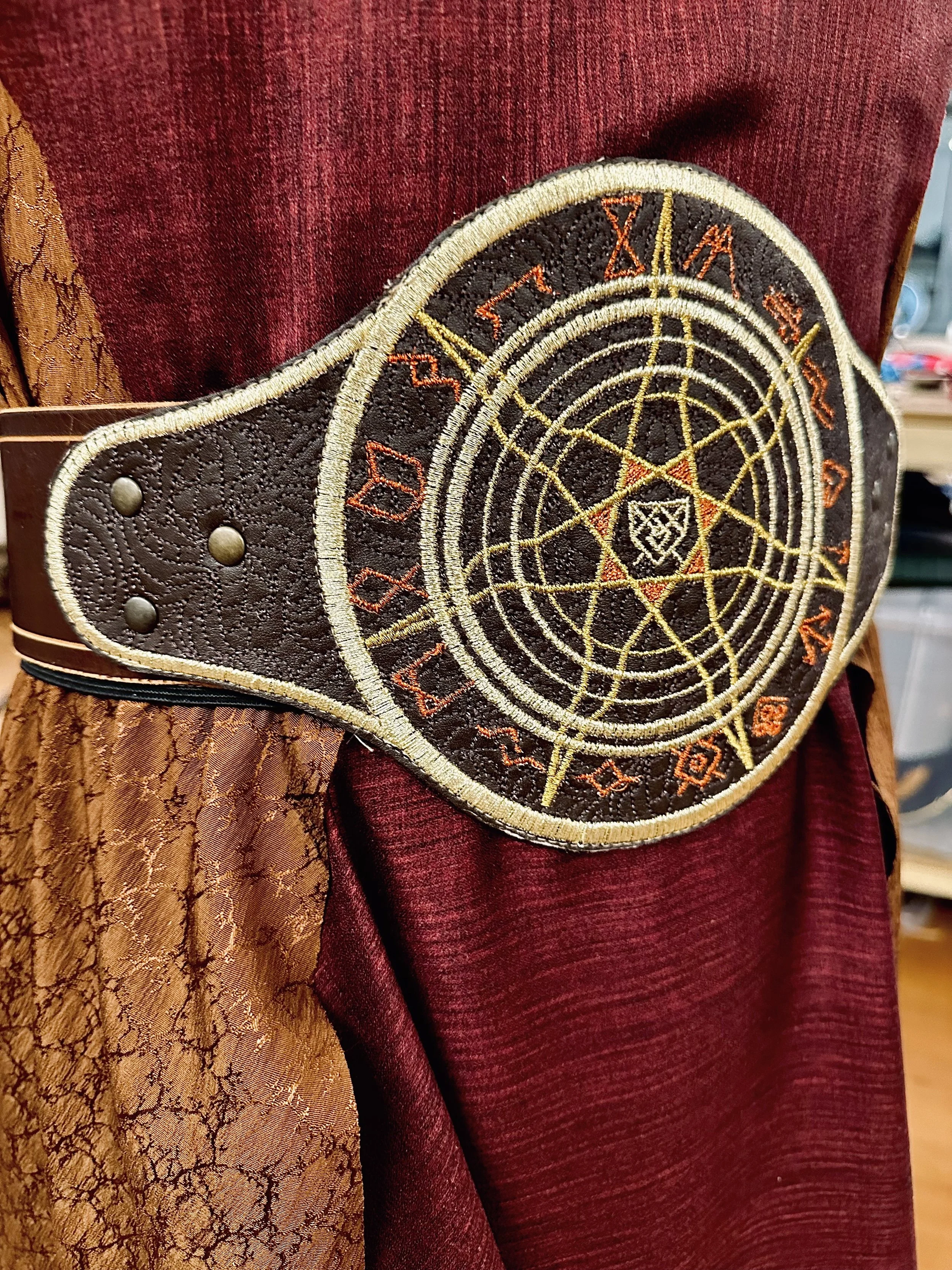 A close-up of a decorative or cosplay belt with a circular embroidered design featuring a pentagram and runes, worn around a person wearing a mix of maroon and textured orange-brown clothing.
