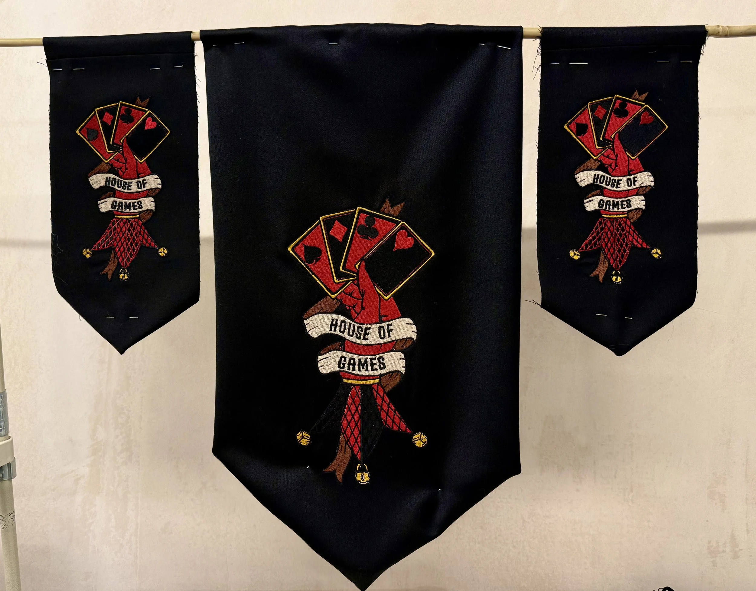 Black banner with embroidered emblem of a jester's hat with playing cards and the words 'House of Games'.