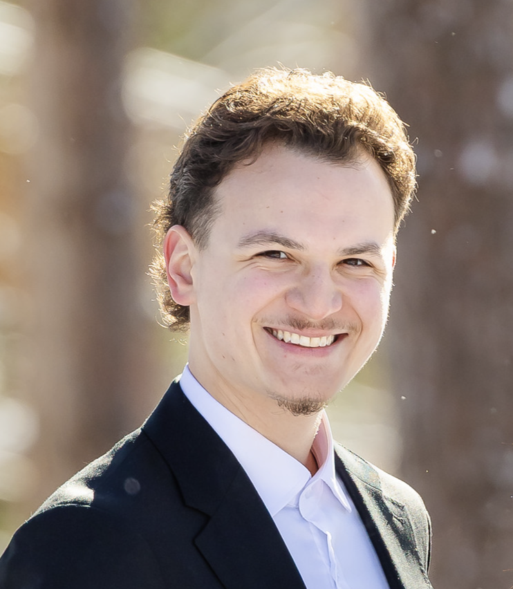 A young man with short, dark curly hair, wearing a suit jacket and white shirt, smiling outdoors on a sunny day.