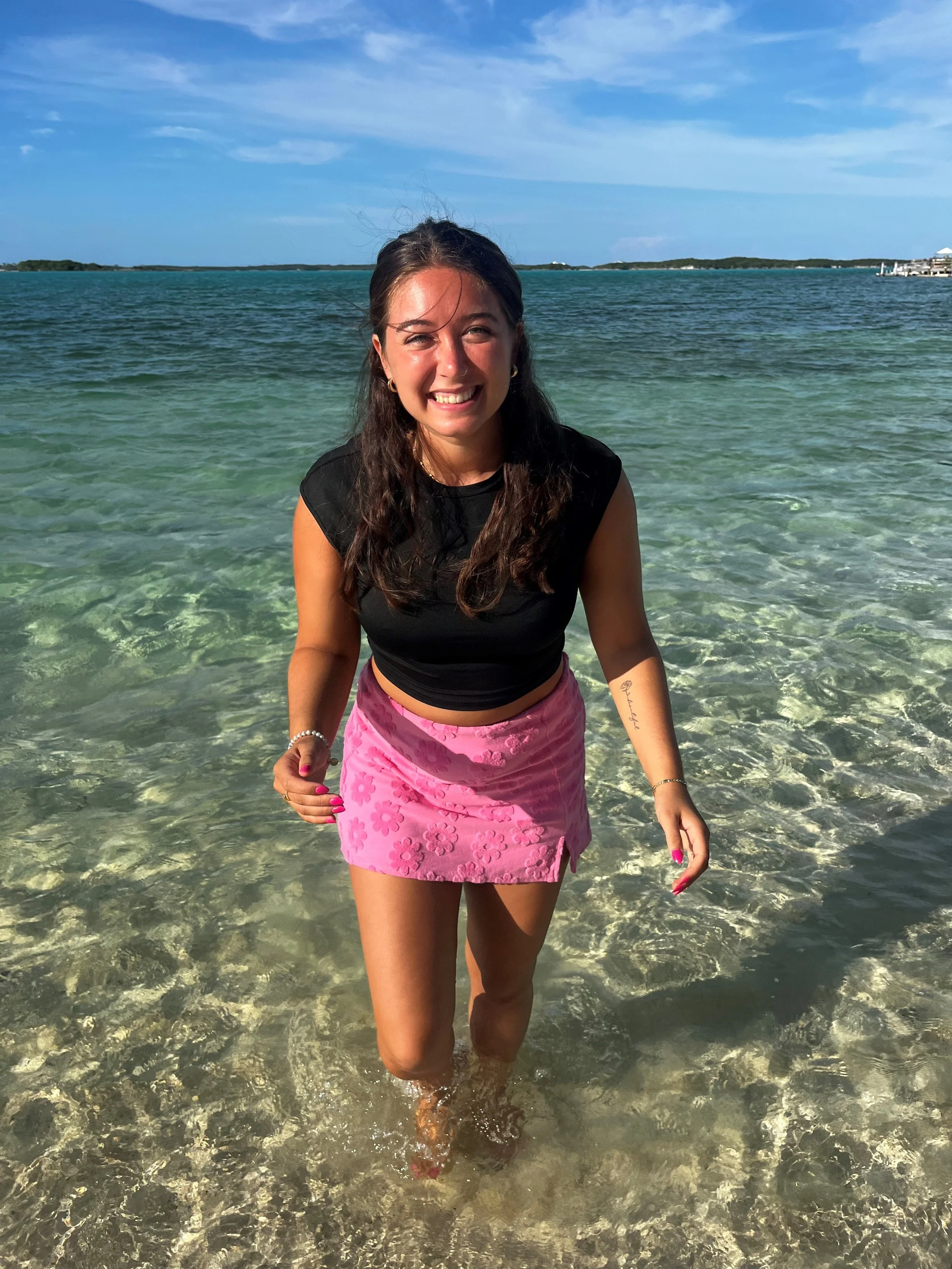 Woman smiling, wearing black top and pink floral skirt, standing in shallow clear water at the beach with a blue sky and distant islands in the background.