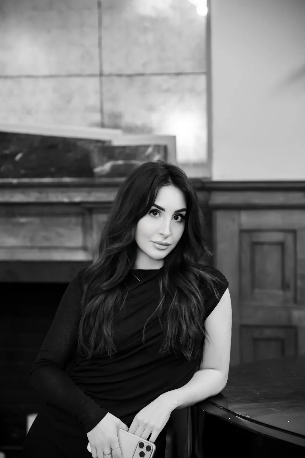 Black and white photo of a woman with long wavy hair, wearing a black dress, holding a smartphone, sitting at a table in a room with wooden paneling.