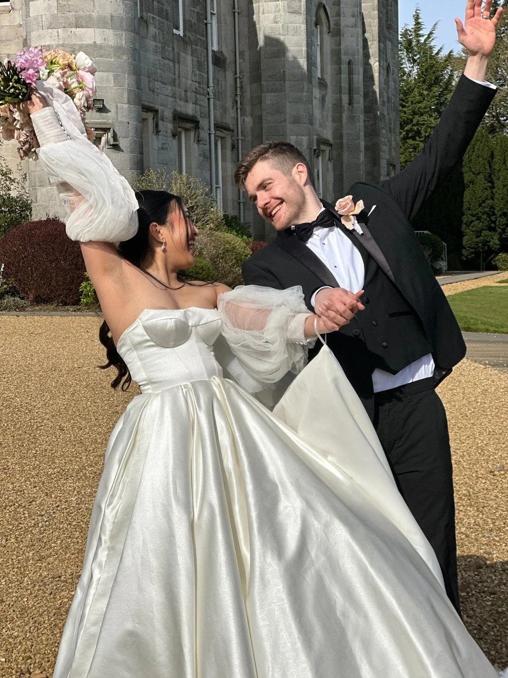 A bride and groom happily dancing outdoors on their wedding day, with the bride holding a bouquet, smiling at each other, and holding hands.