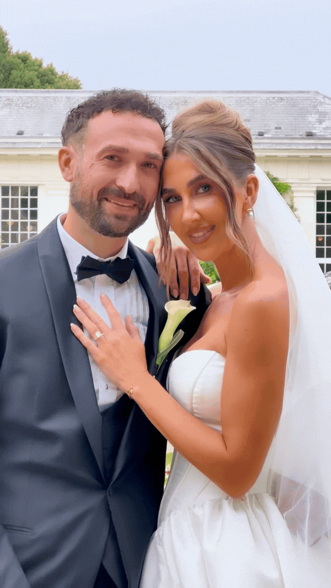 A newlywed couple outdoors, the groom in a tuxedo with a black bow tie, and the bride in a strapless white wedding dress with a veil, standing closely together and smiling at the camera.