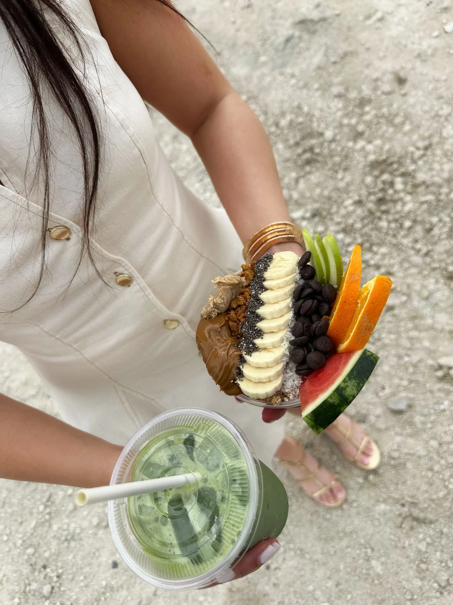 A woman in a cream-colored dress holding a bowl of colorful fruit and dessert pieces, and a cup of green juice with a straw, standing on a gravel surface.