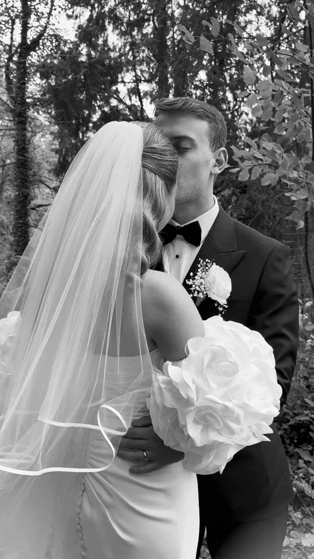 A bride and groom sharing a kiss outdoors, with the bride holding a large bouquet of flowers and wearing a veil, and the groom dressed in a tuxedo with a bow tie and boutonniere, surrounded by trees and foliage.