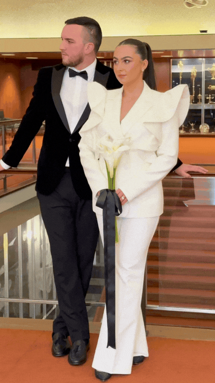 Man and woman dressed in formal attire standing together at a hotel or event venue. The man is wearing a tuxedo, and the woman is wearing a white suit and holding a white calla lily.