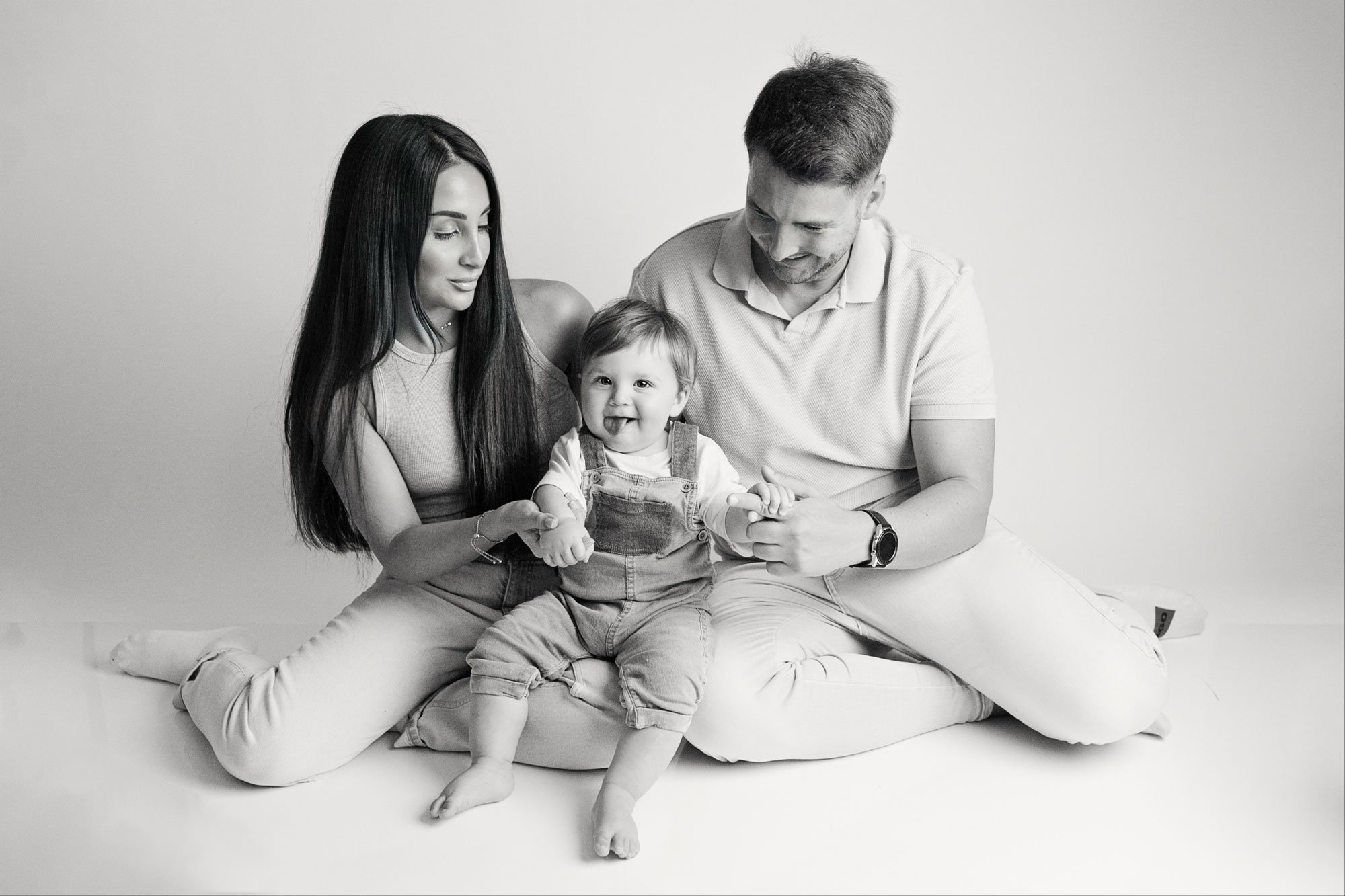A black and white photo of a family of three sitting on the floor. The mother has long dark hair and is holding the child's hand. The father with short hair is playing with the child. The child, a toddler with short hair, is sitting between the parents, smiling, with their tongue out, wearing overalls and a t-shirt.