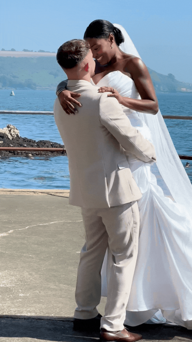 A couple dancing outdoors near a body of water during a wedding, with the woman wearing a white wedding dress and the man in a beige suit.