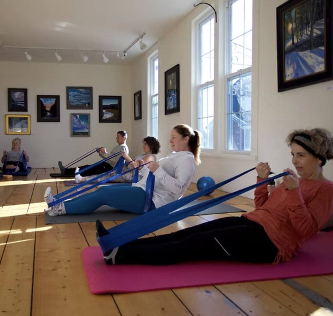 Women participating in a yoga or stretching class using resistance bands in a bright studio with wooden floors, artwork on the walls, and natural light from large windows.