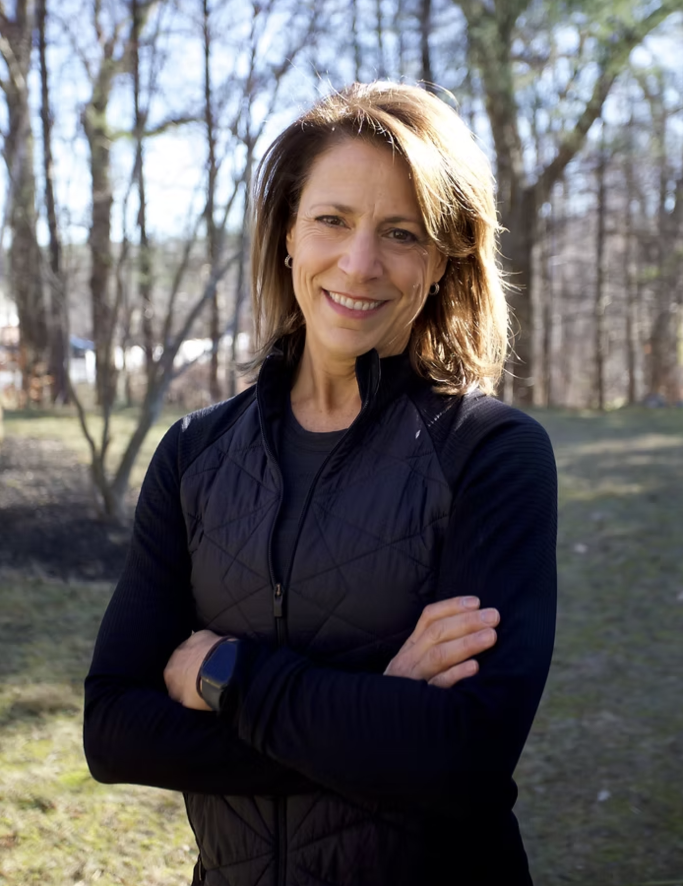 A woman with light brown hair smiling outdoors in a wooded area, wearing a black athletic jacket and a smartwatch on her left wrist.