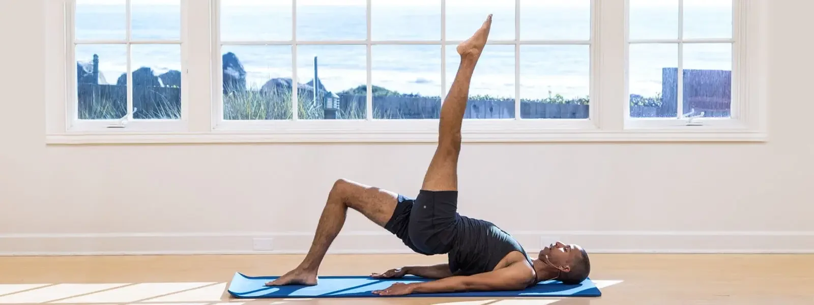 A person doing a yoga pose in a brightly lit room with large windows showing a view of rocks and greenery outside.