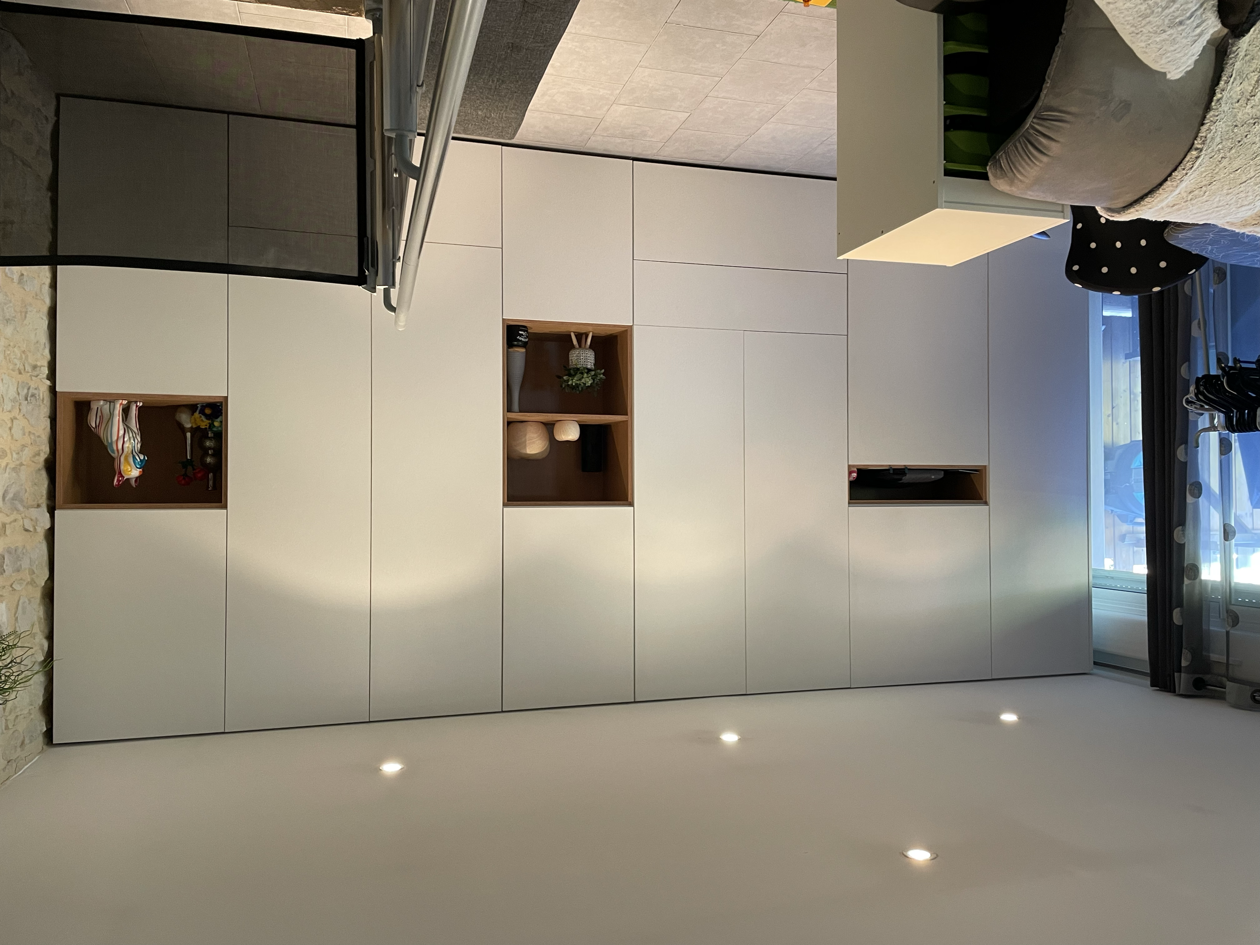 Garde-robe blanche avec des niches en bois, dans un salon moderne, à côté d'un canapé gris, avec fenêtre avec rideaux à pois et une table basse.