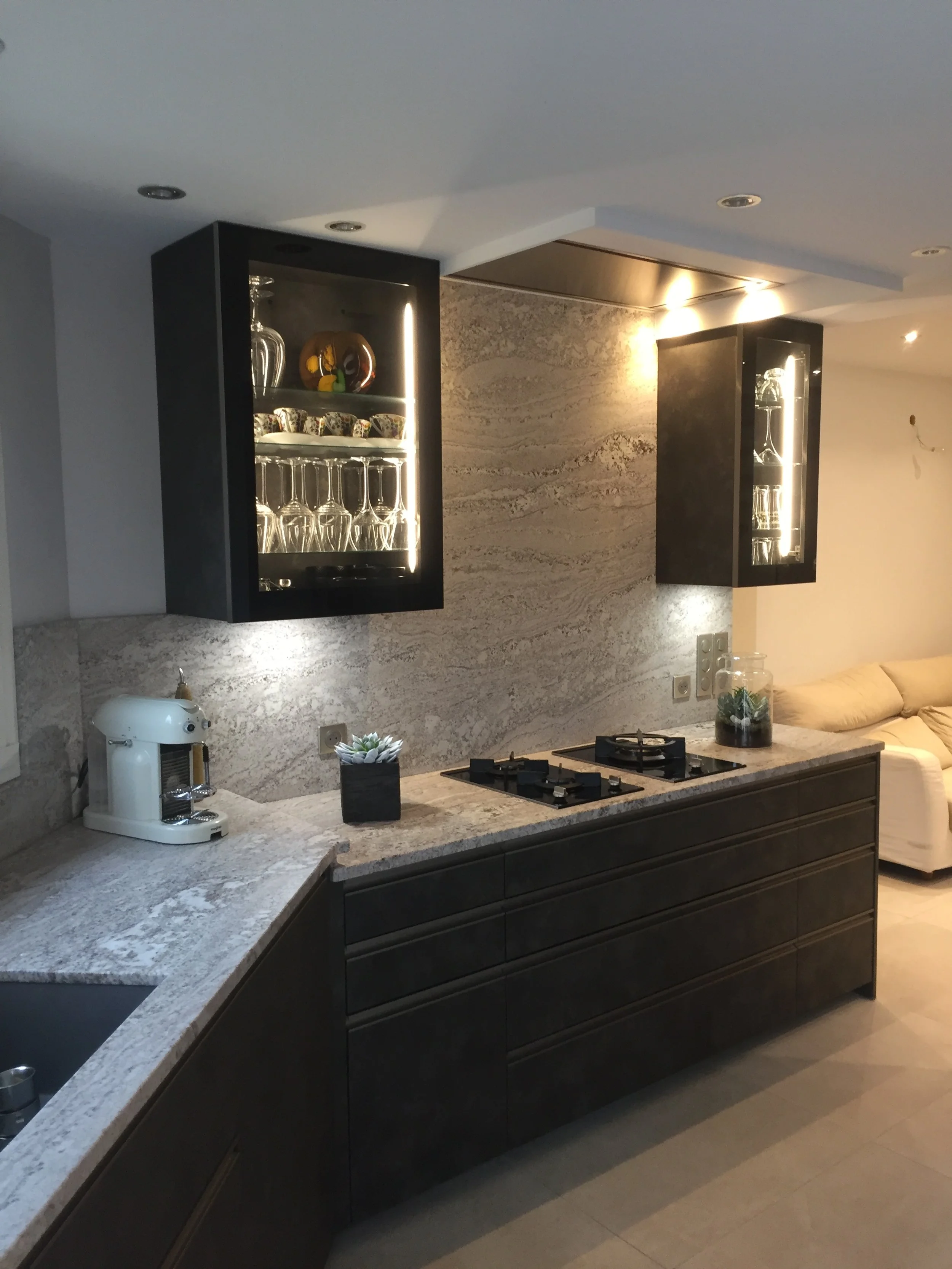 Une cuisine moderne avec des armoires noires, un plan de travail en pierre, une cafetière blanche, et un mur en pierre naturelle. Deux armoires murales avec éclairage intégré contenant de la vaisselle et des verres.