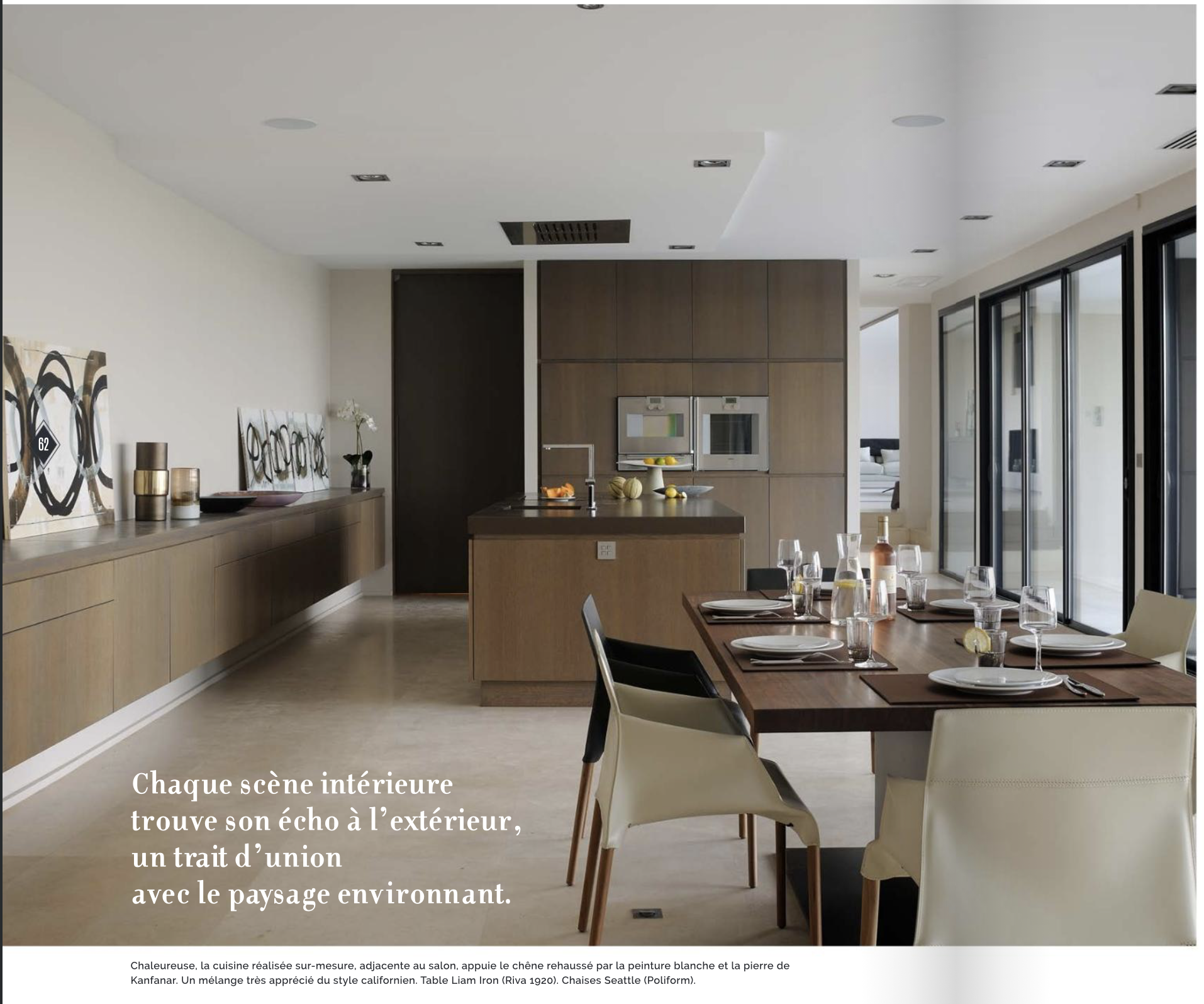 Une salle à manger moderne avec une table en bois entourée de chaises, une cuisine en arrière-plan avec un îlot et des appareils encastrés, grands panneaux coulissants en verre à droite, décorations et vaisselle sur la table. Le texte en français mentionne le lien entre intérieur et extérieur, le paysage environnant, et décrit les matériaux et le style de la cuisine.