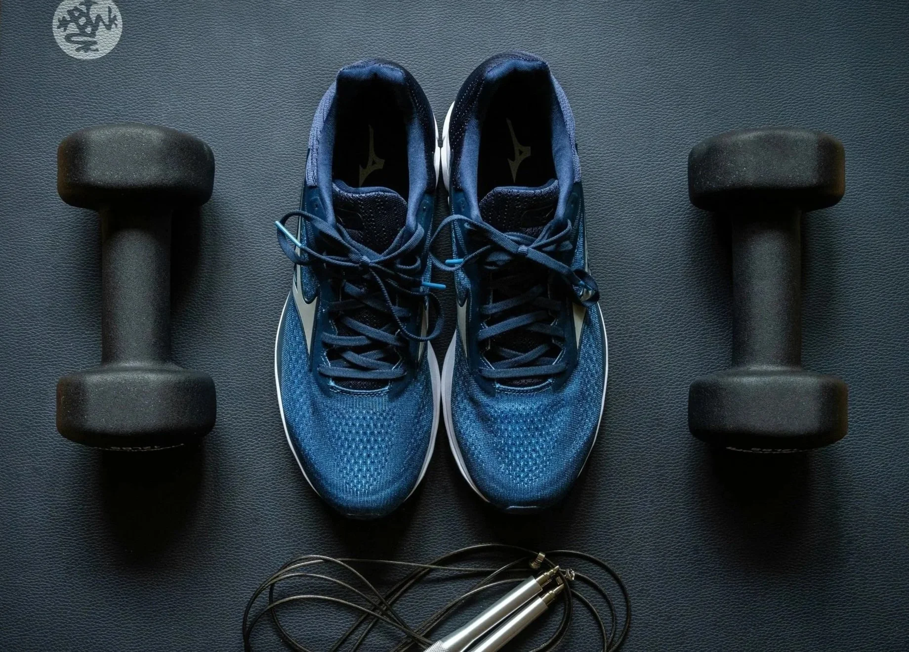 A pair of blue athletic shoes, two black dumbbells, and a measuring tape on a dark textured surface, with an exercise logo in the top left corner.