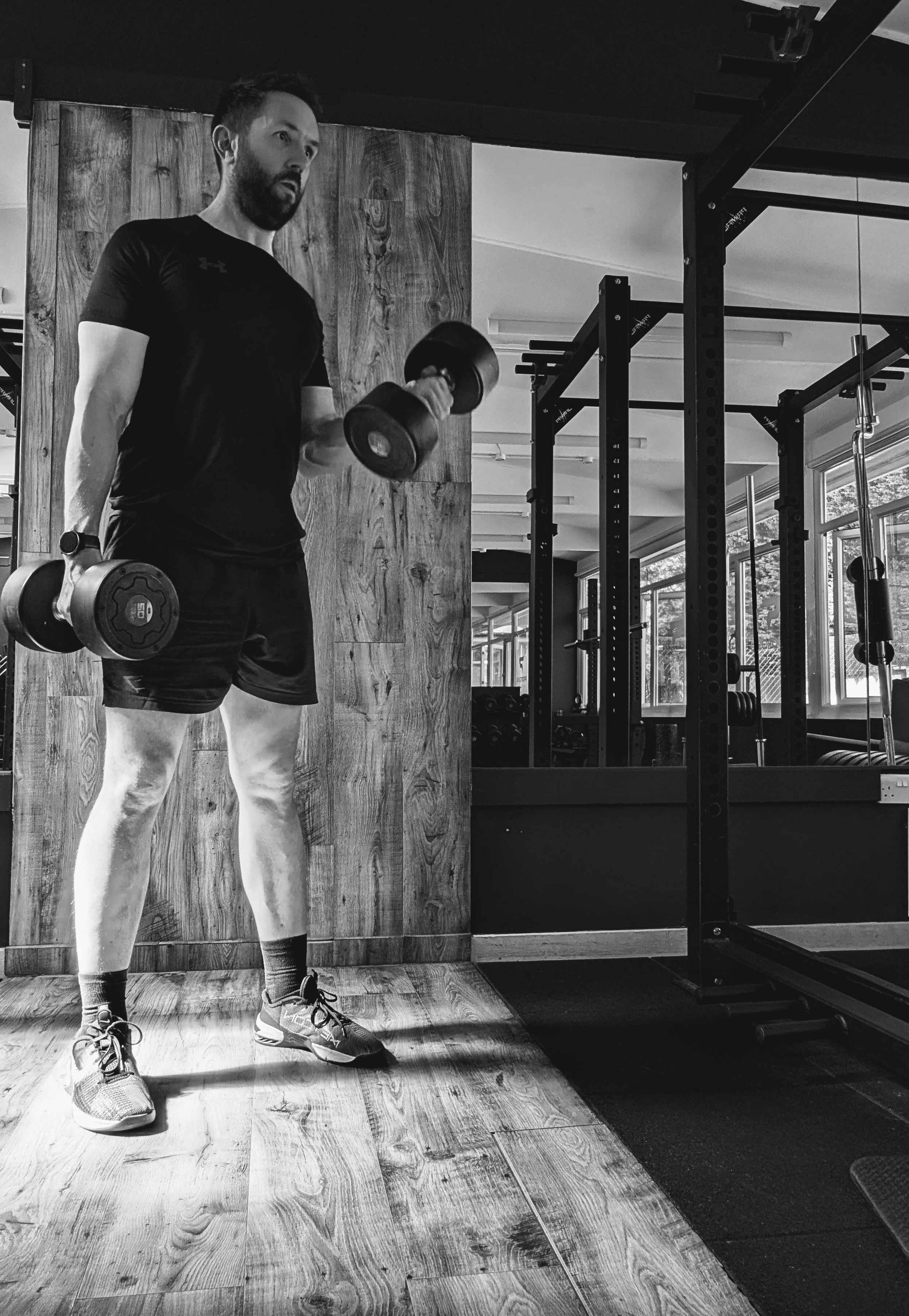 A personal trainer based in Chobham and Surrey lifting a dumbbell in a gym, wearing a black t-shirt, shorts, and running shoes, with gym equipment and large windows in the background.