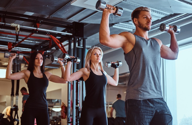 A personal trainer based in Chobham and Surrey. Three people working out with dumbbells in a gym, with a man in the foreground and two women behind him.