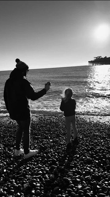 A personal trainer based in Chobham and Surrey. A dad with his daughter that understands life is a balancing act. He stands on a pebble beach near the water's edge during sunset, with a pier in the background.