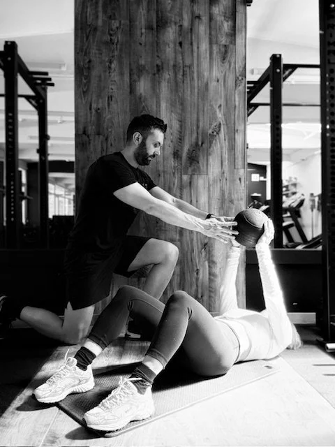Personal trainer in Chobham and Surrey helping a woman exercising in a gym. The woman is lying on a mat, holding a medicine ball, while the man is assisting her in a workout. The gym has weight racks and equipment in the background.