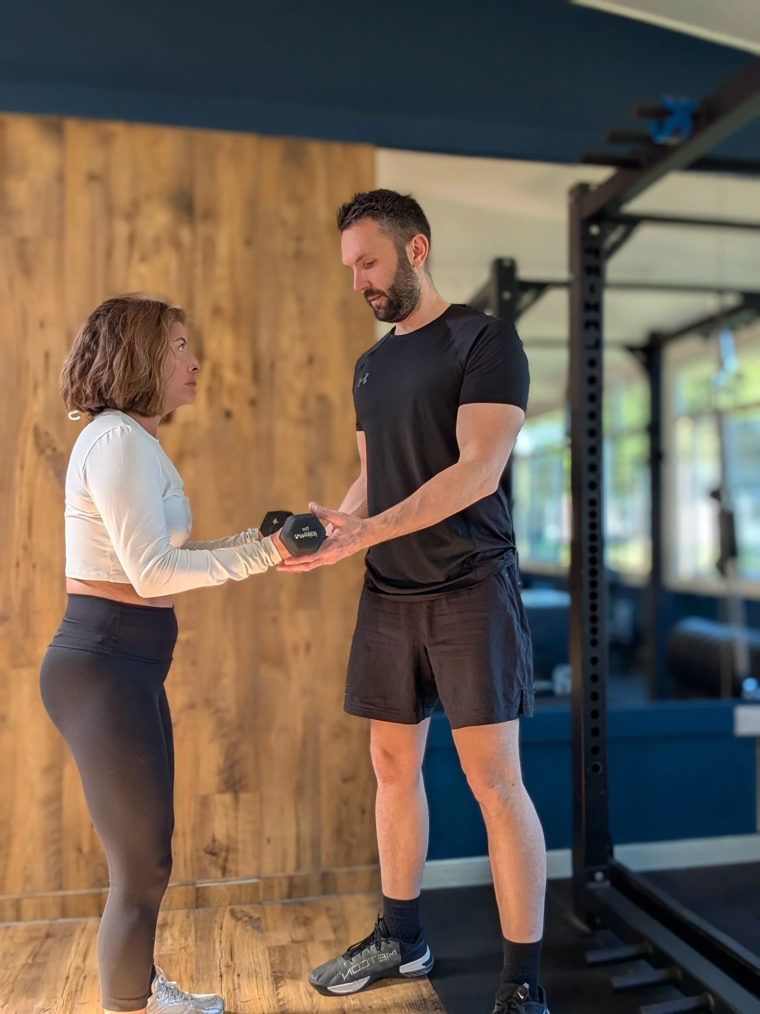 A personal trainer based in Chobham and SurreyA woman and a man in a gym. The woman, holds a dumbbell. The man,  is standing in front of her, possibly giving instructions or discussing. The gym has wooden flooring and a wooden wall in the background.