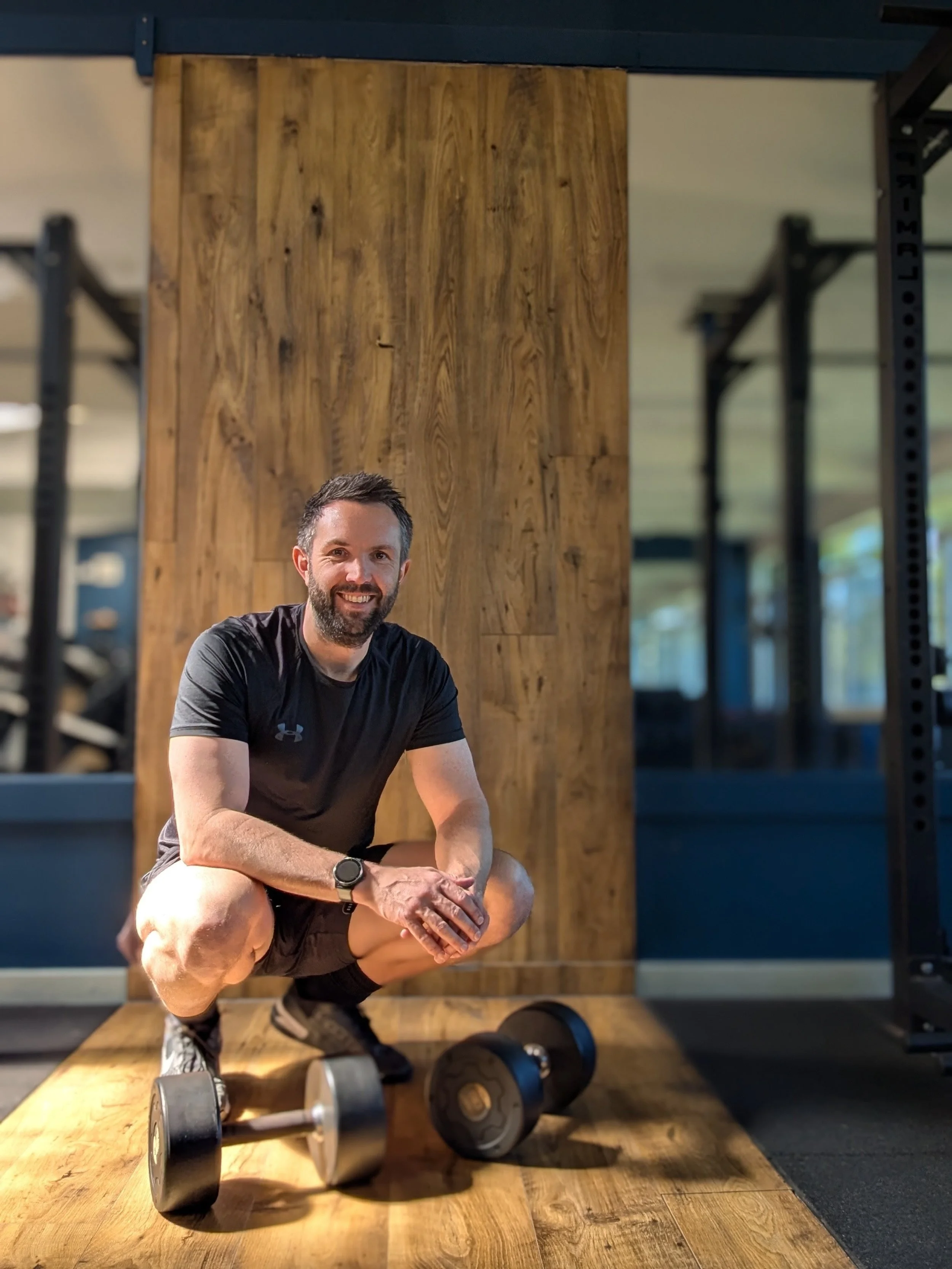 A personal trainer based in Chobham and Surrey in workout clothes sitting on a wooden bench in a gym, surrounded by dumbbells, with a wooden wall and exercise equipment in the background.