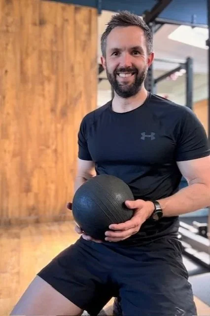 A personal trainer based in Chobham and Surrey smiling in the gym, holding a medicine ball, wearing a black athletic shirt and shorts.