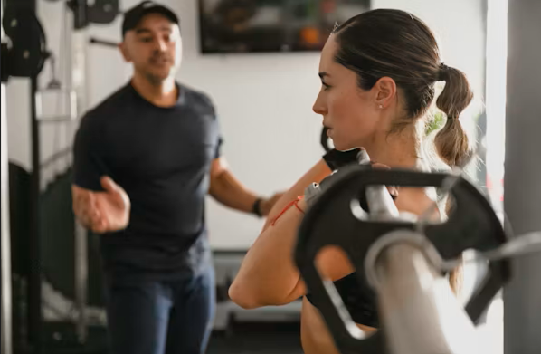 A woman receiving instructions from a trainer at a gym with weights and fitness equipment.