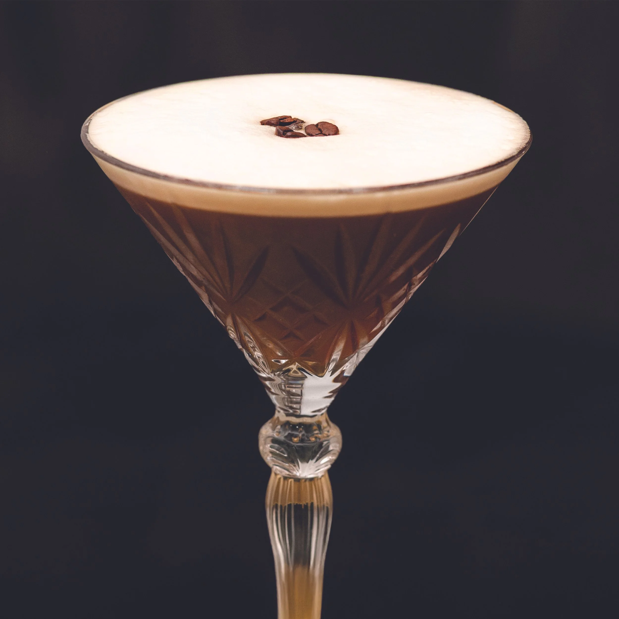 A crystal glass with a geometric pattern filled with a dark brown cocktail, topped with a white foam and a few coffee beans on top.