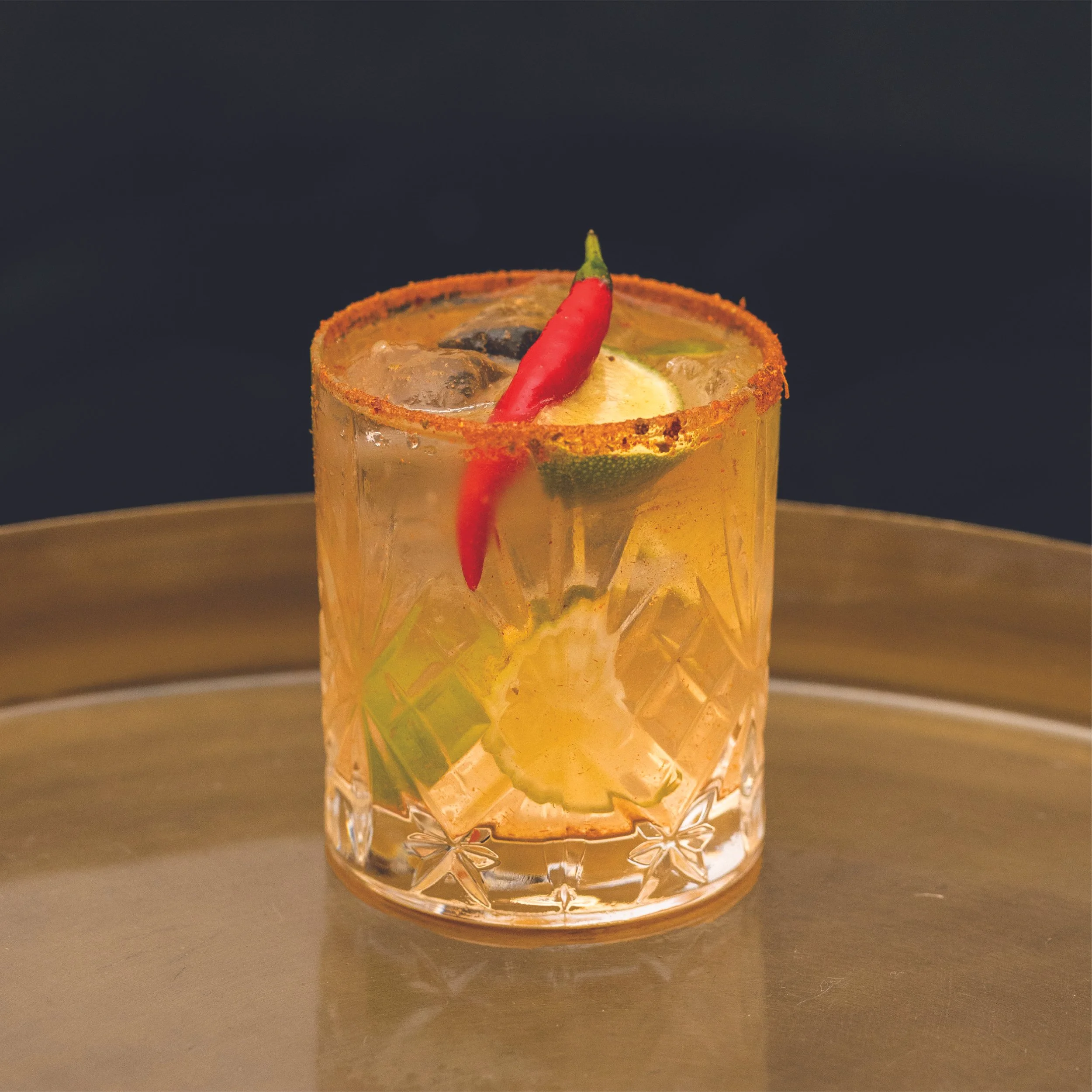 A glass of tequila cocktail with lime, lemon slices, a red chili pepper, and salt rim, on a gold tray.