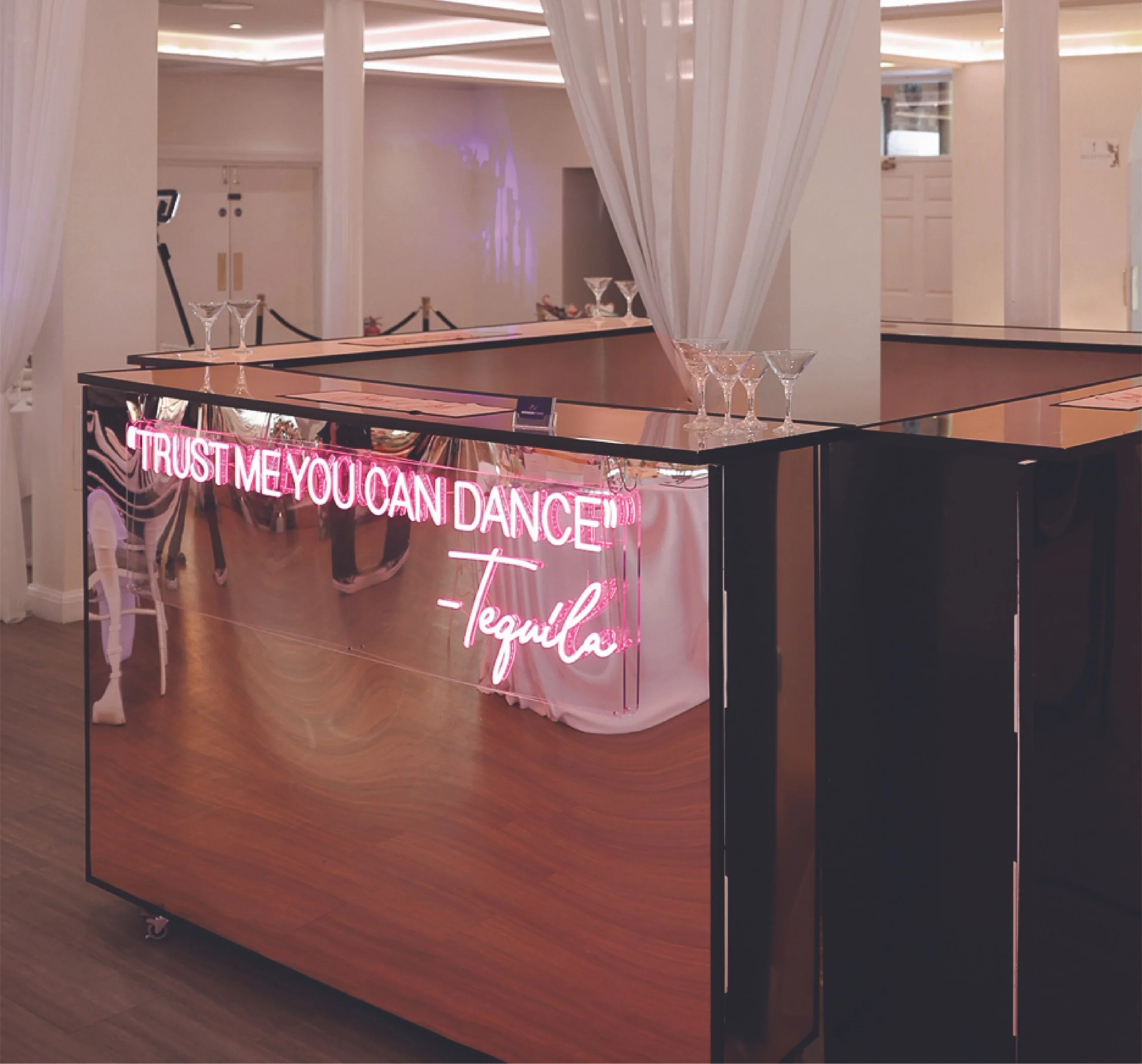 A bar or counter with a pink neon sign that reads, 'Trust me, you can dance,' followed by 'Tequila.' The bar has a reflective surface, and there are empty cocktail glasses on top. In the background, white curtains and a soft indoor lighting create an elegant atmosphere.