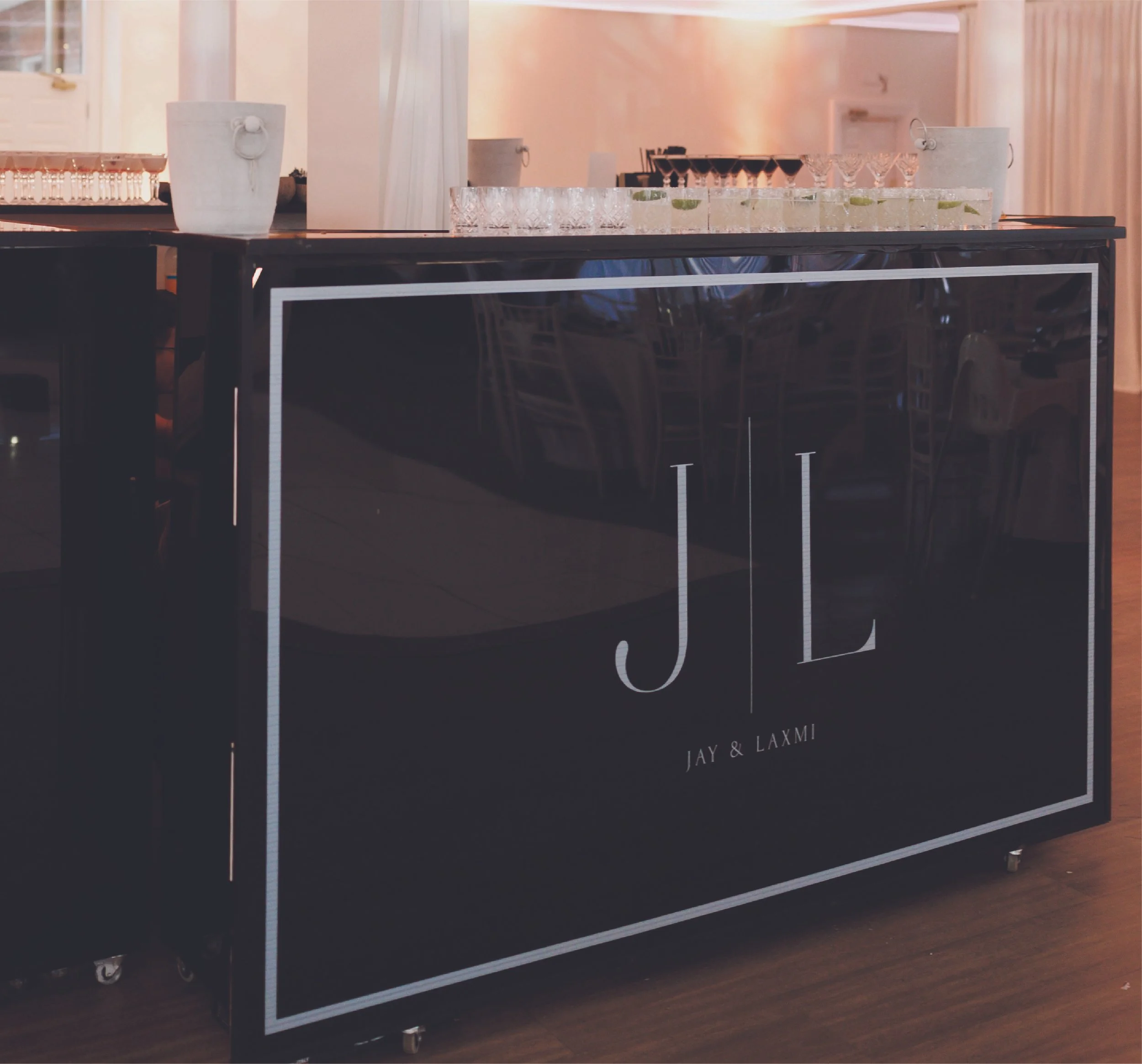 A black bar or drink station with the initials 'J L' and the names 'Jay & Laxmi' below, situated in a decorated indoor setting with glasses and drinks on top.