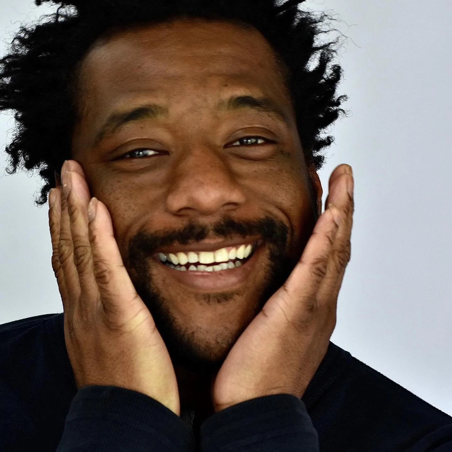 A man smiling broadly with his hands on his cheeks, looking joyful with a clear complexion and short, curly hair.