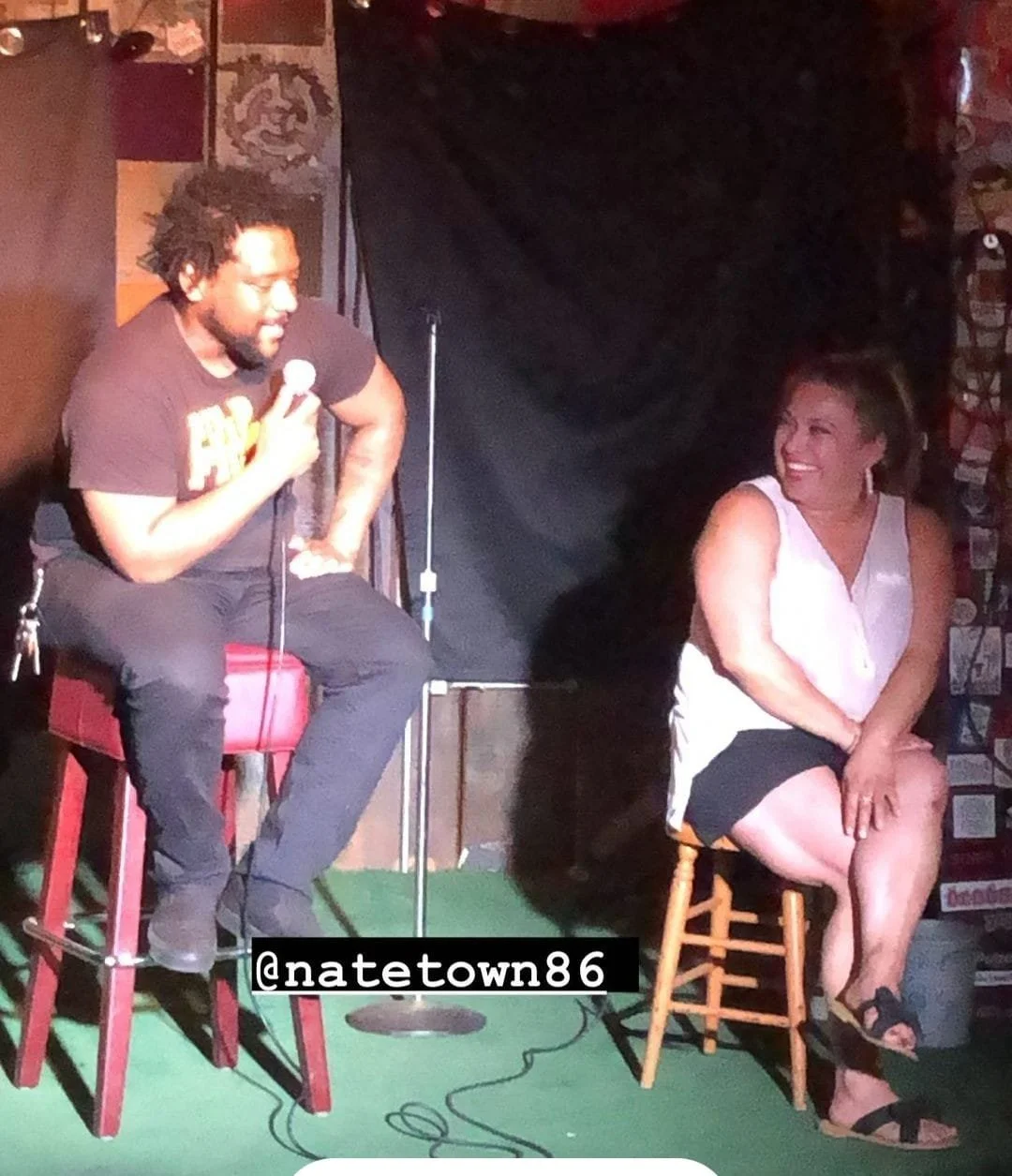 A man and a woman sitting on stools on a small stage, engaging in a comedic or conversational act in a dimly lit venue. The man is holding a microphone and leaning towards the woman, who is smiling and looking at him. The background features a black 