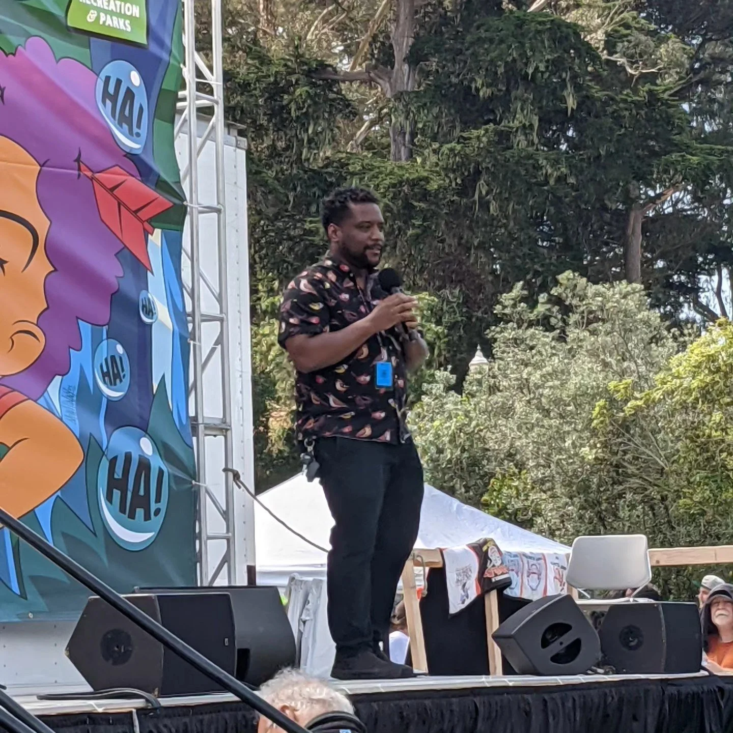 A man with a beard and hairstyle standing on an outdoor stage, holding a microphone, with a colorful banner featuring a cartoon character and speech bubbles behind him, surrounded by trees.