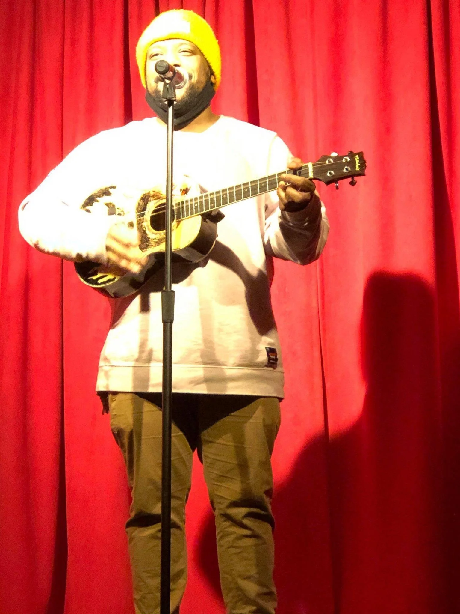 A person wearing a yellow knit cap, black face mask, and white sweatshirt, singing into a microphone and playing an acoustic guitar on stage with red curtains in the background.