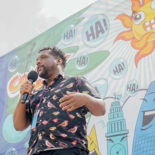 Man speaking into a microphone in front of a colorful comic-style backdrop with the sun and speech bubbles saying 'HA!'