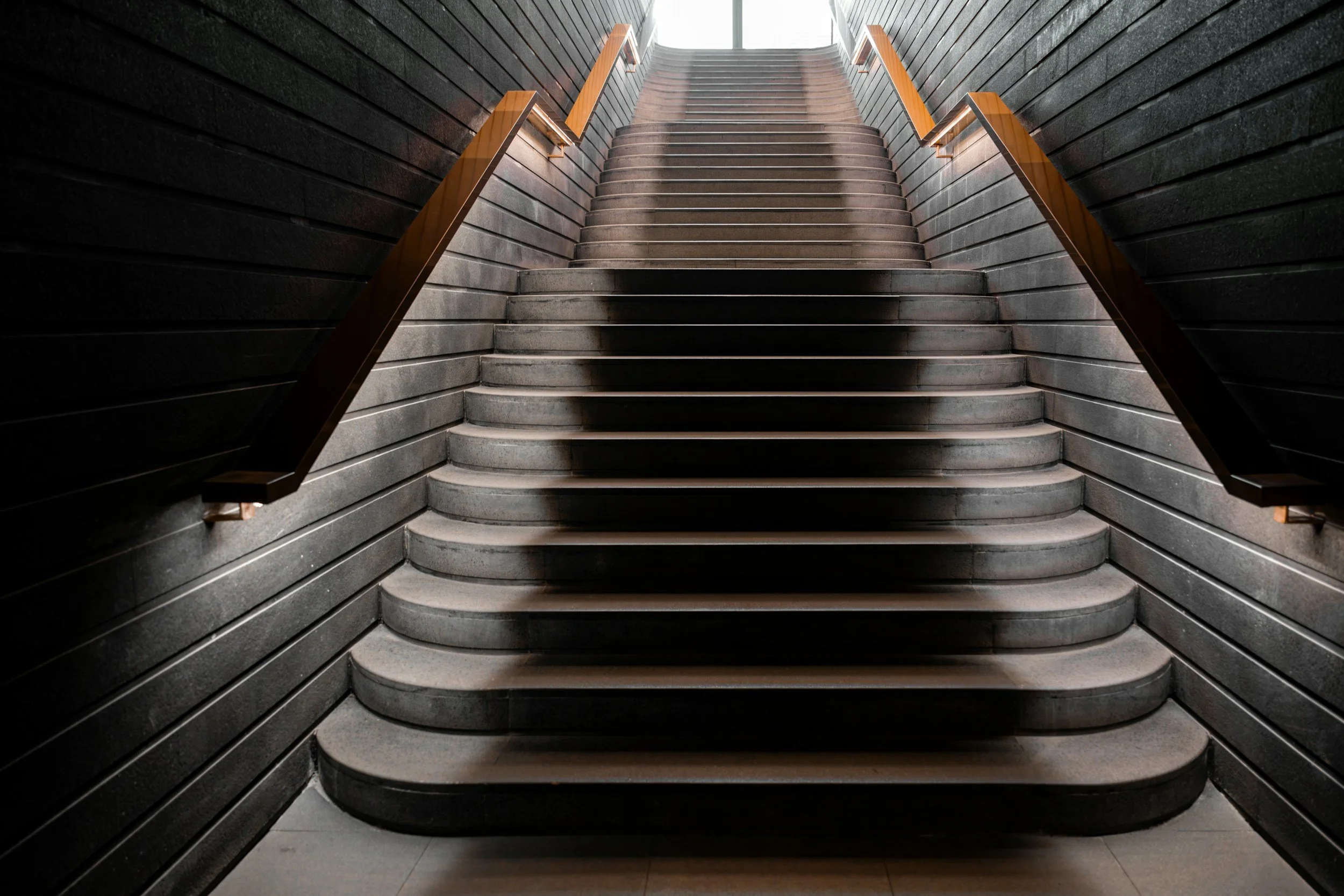 Indoor staircase with dark wood handrails and black walls, leading upward toward a light source.