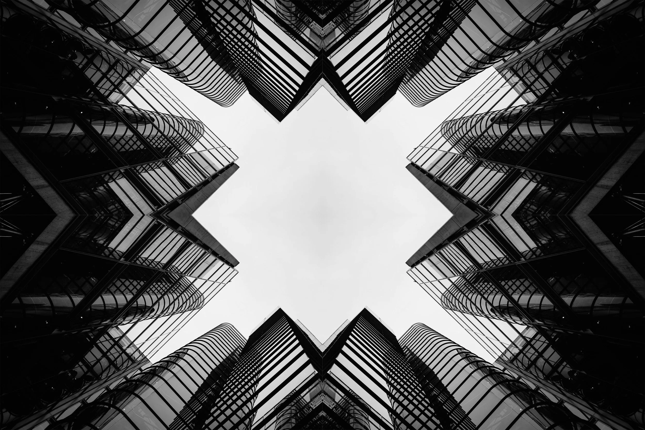 Black and white photograph of modern skyscrapers taken from below, looking up at the sky, creating a symmetrical star-like pattern.