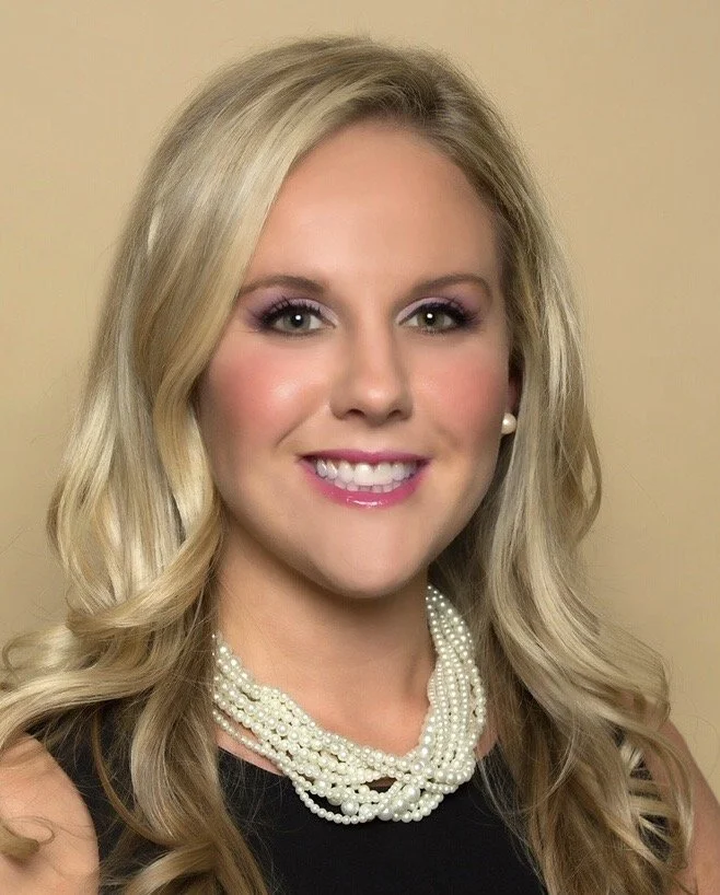 A woman with long, wavy blonde hair smiling, wearing makeup with purple eyeshadow, pearl earrings, and a multi-strand pearl necklace, against a beige background.
