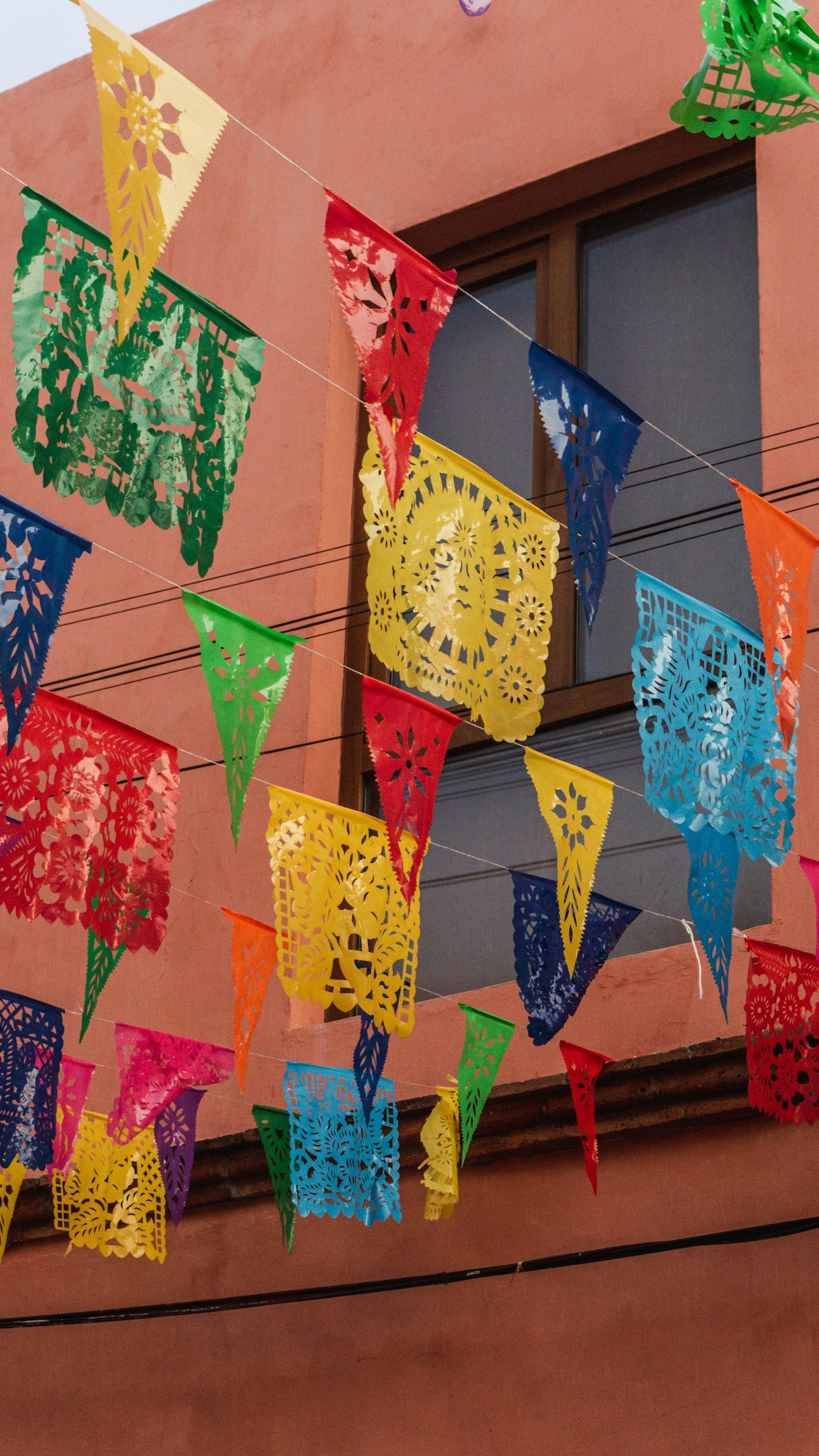 Colorful papel picado banners hanging in the air outdoors against a pink building with brown window frames.