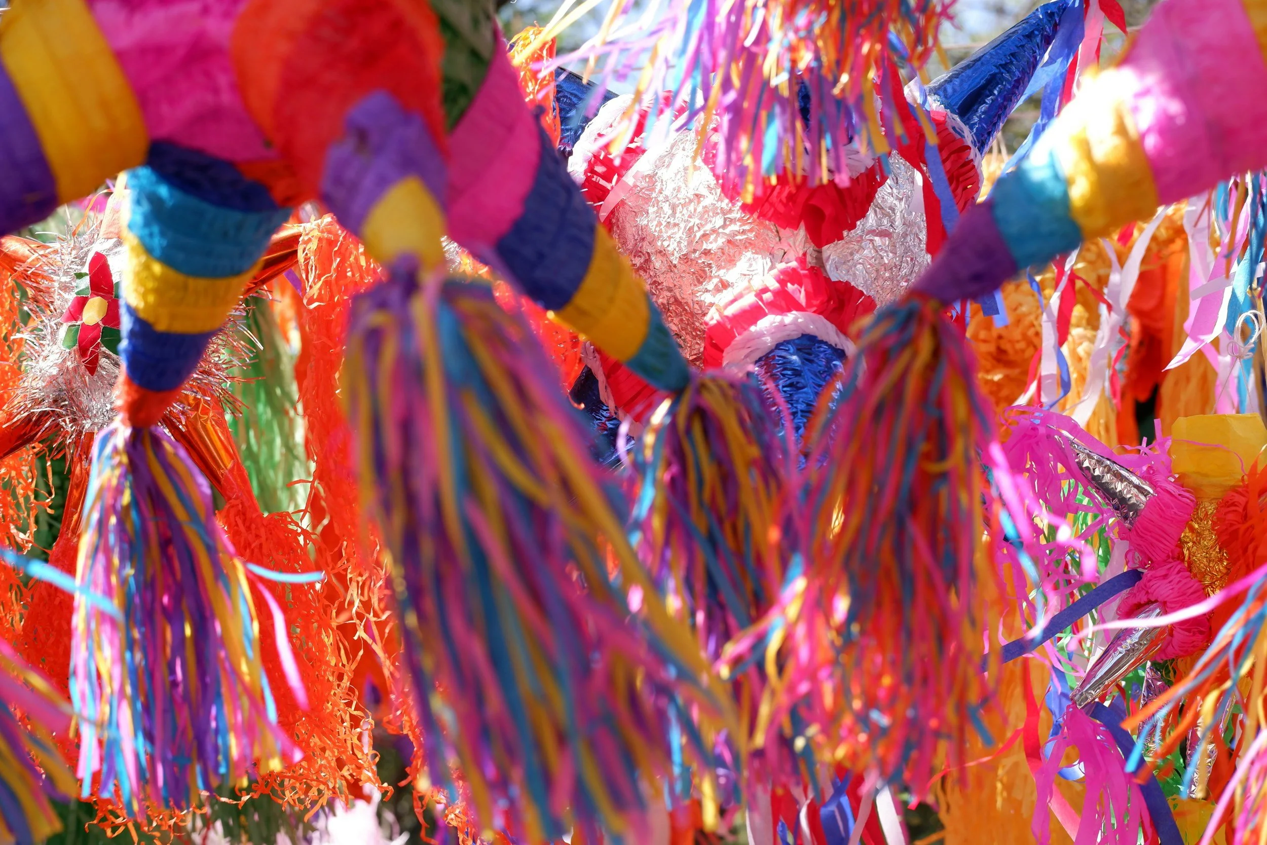 Colorful papel picado decorations with fringed, shiny, and braided paper strips in pink, orange, blue, purple, yellow, and metallic colors hanging in the air.