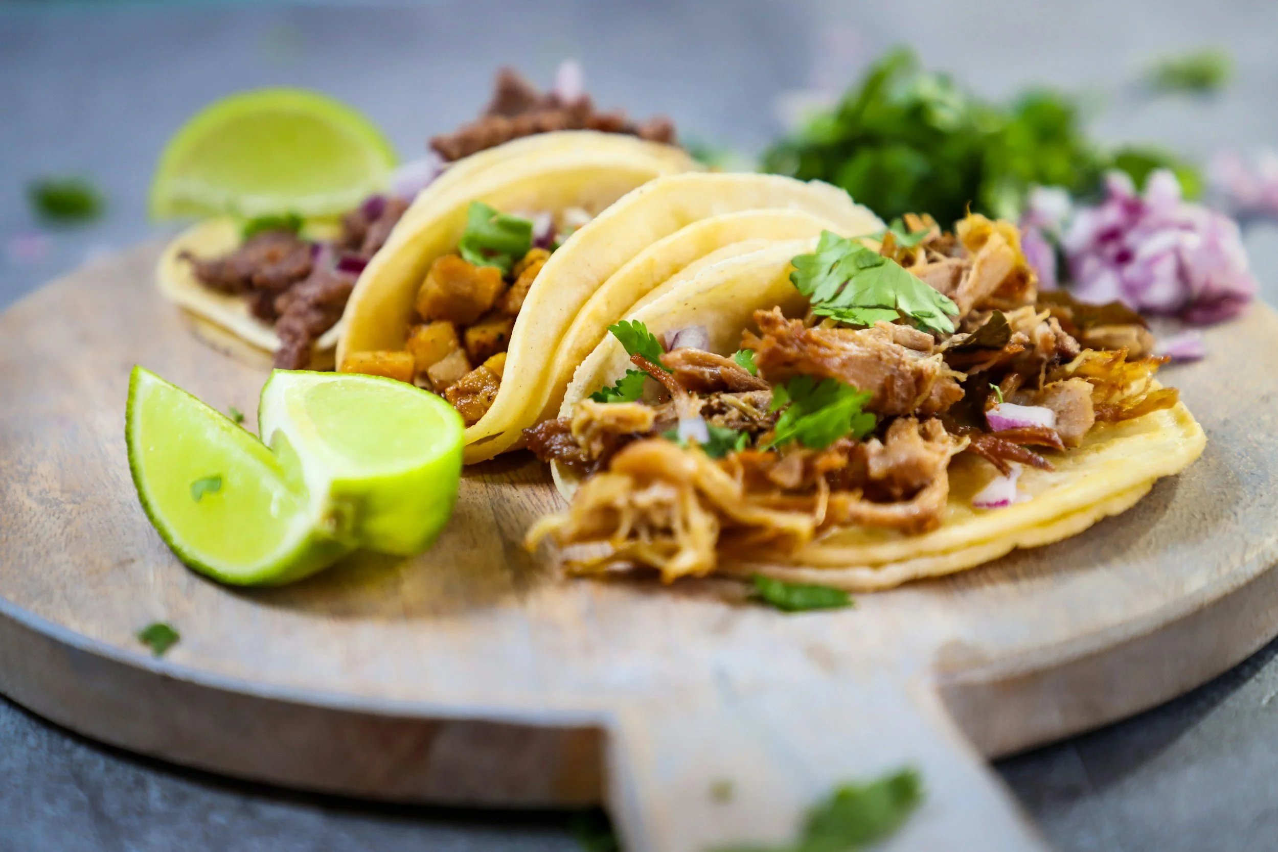 Three tacos filled with shredded pork, chopped onions, cilantro, and served with lime wedges on a wooden platter.