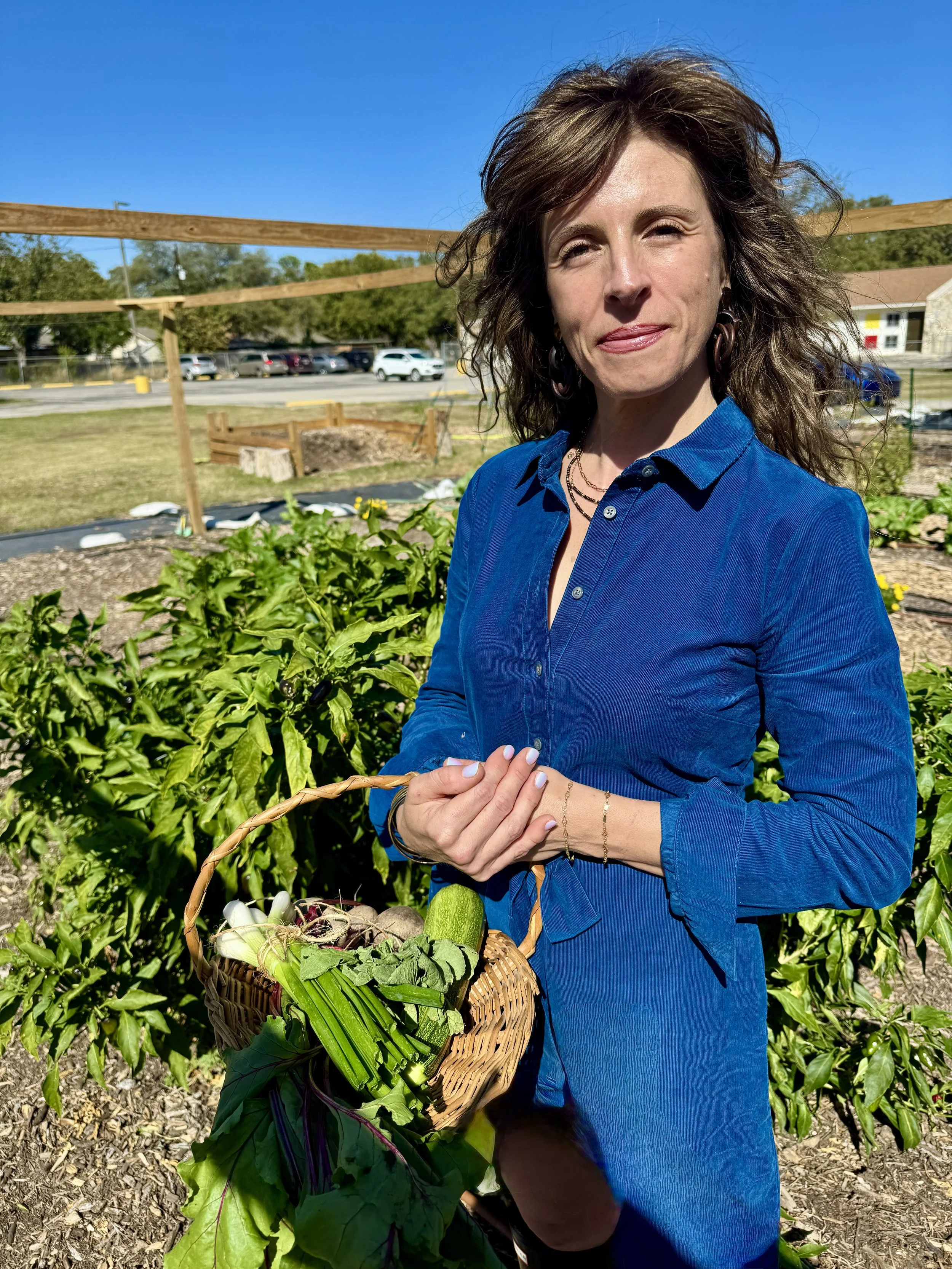 A woman in a blue button-up shirt holding a basket of freshly harvested vegetables in a garden.