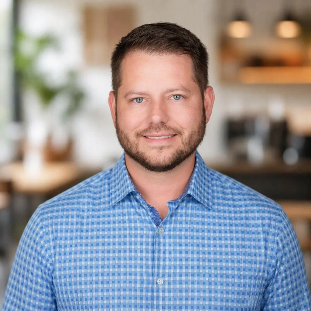 A man with short brown hair, blue eyes, and a beard wearing a blue checkered shirt, smiling in an indoor setting.