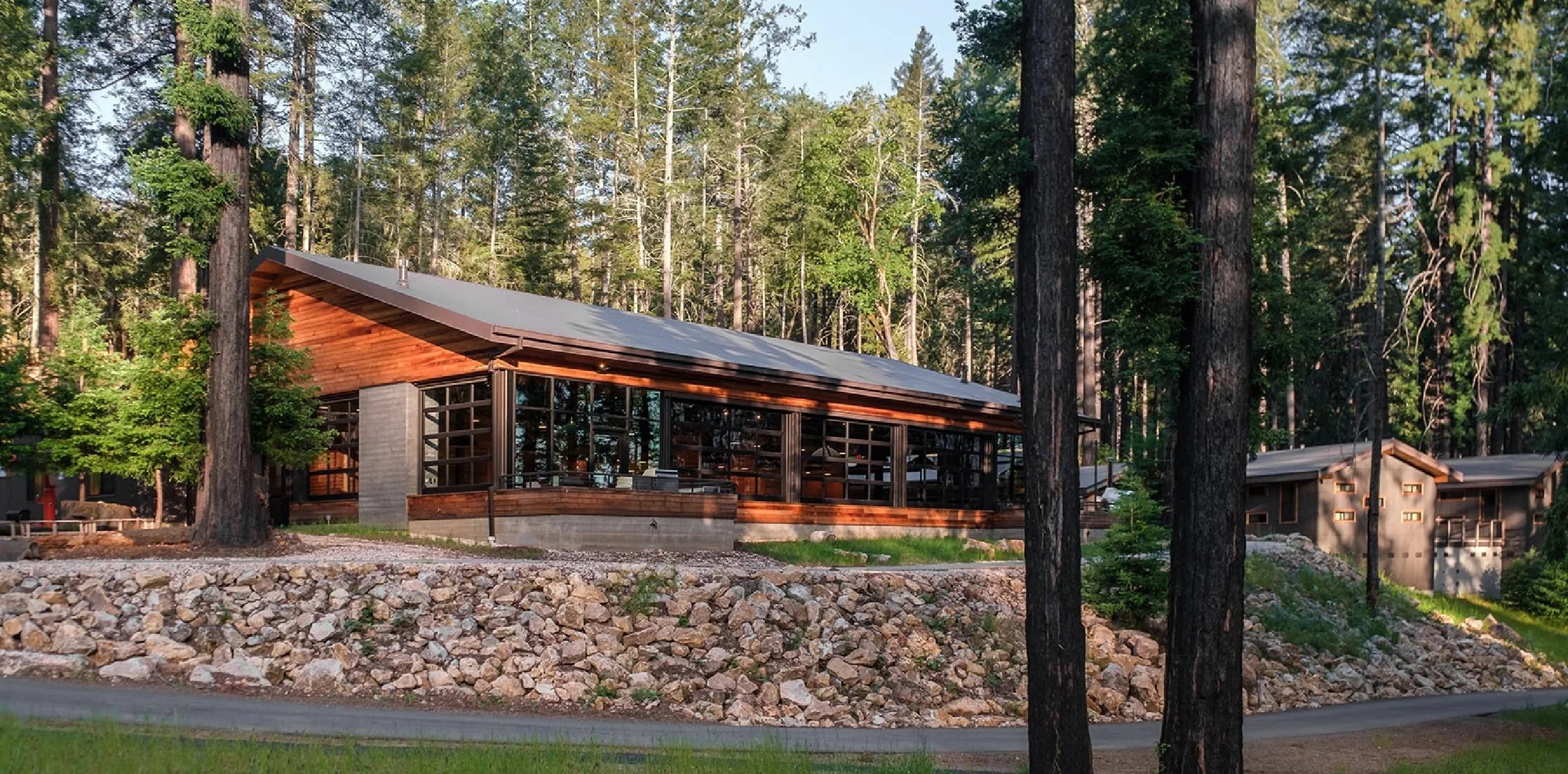 Modern house with large glass windows and a sloped roof, situated in a forested area with tall trees and rocky landscape in the foreground.