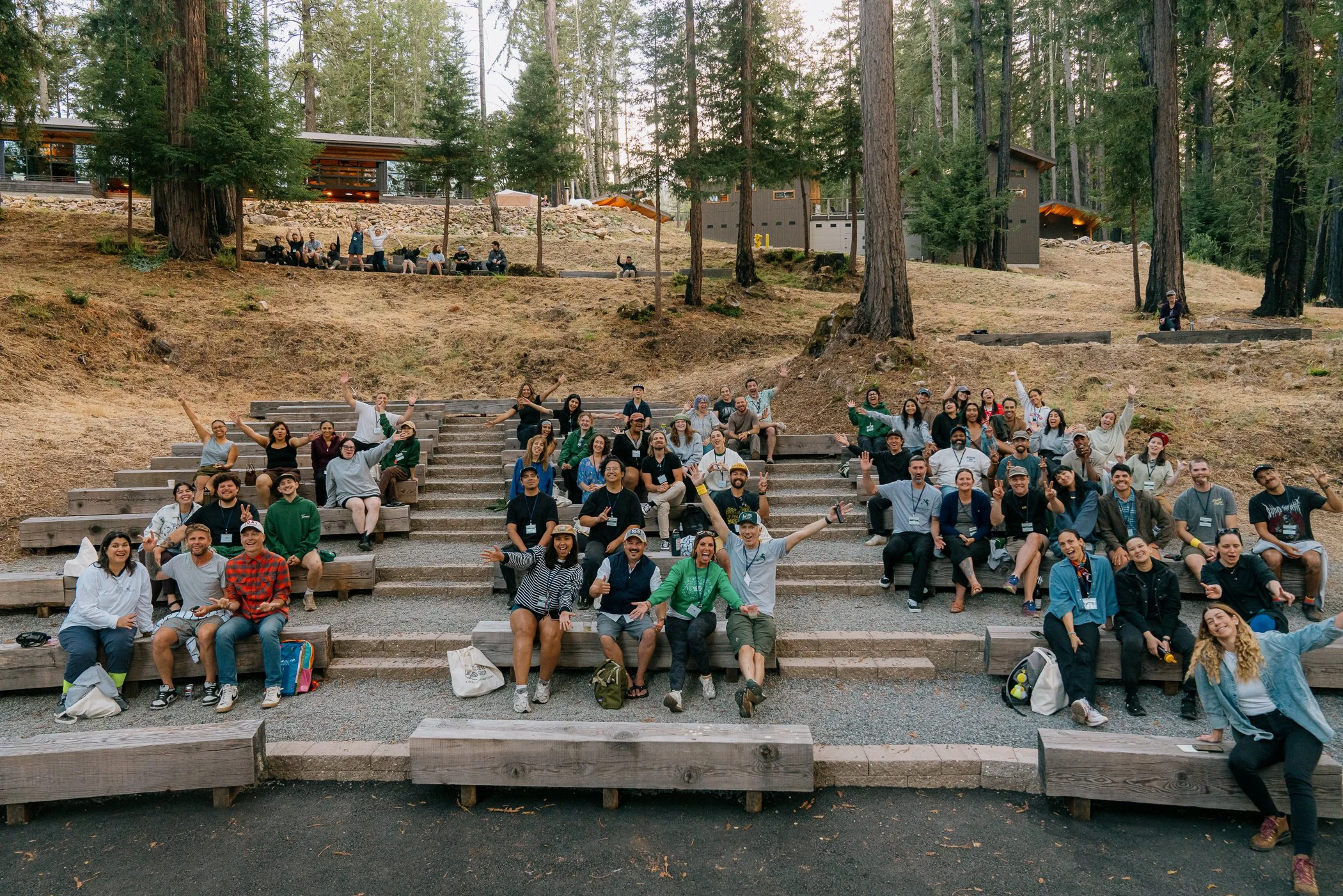 A large group of people sitting and standing on outdoor wooden steps in a forested area, smiling and waving at the camera.