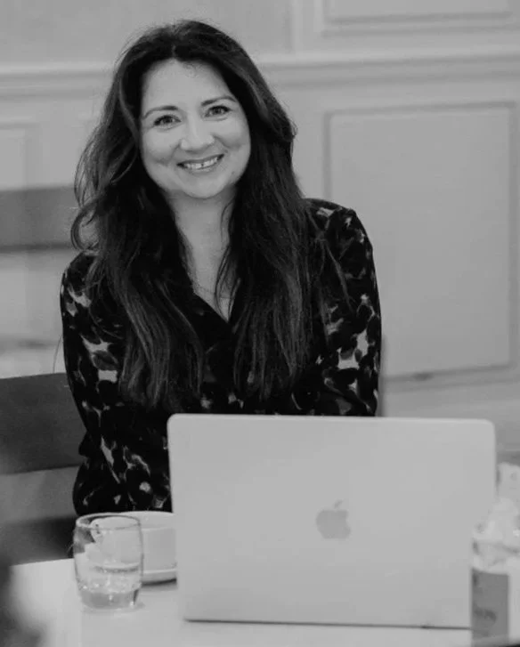 Carla with long dark hair smiling at a table with a laptop and a glass of water in a room with paneled walls.