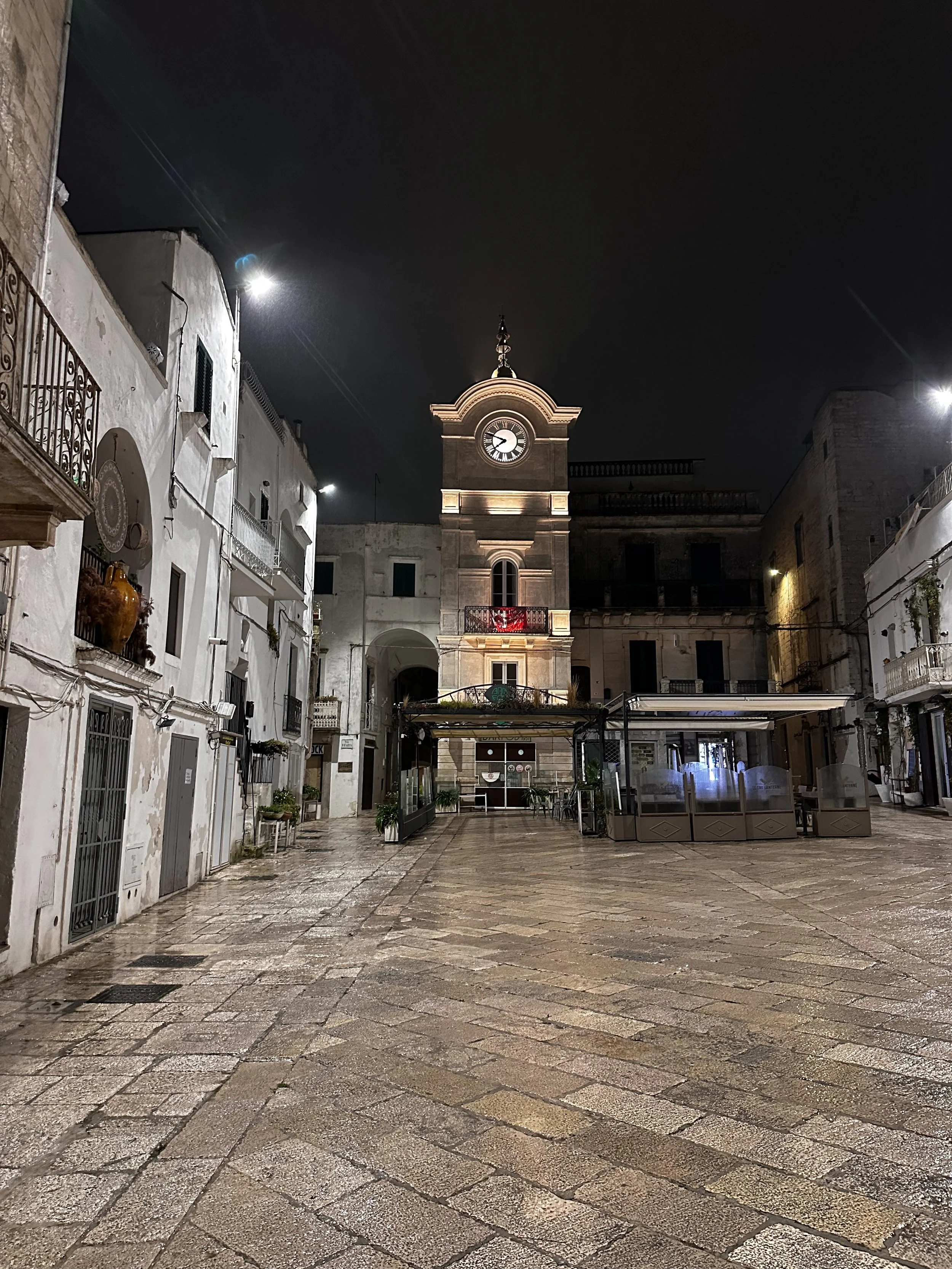Una piazza vuota con pavimento in pietra, un edificio con un orologio sulla torre al centro e alcuni negozi o ristoranti ai lati, illuminata di notte.