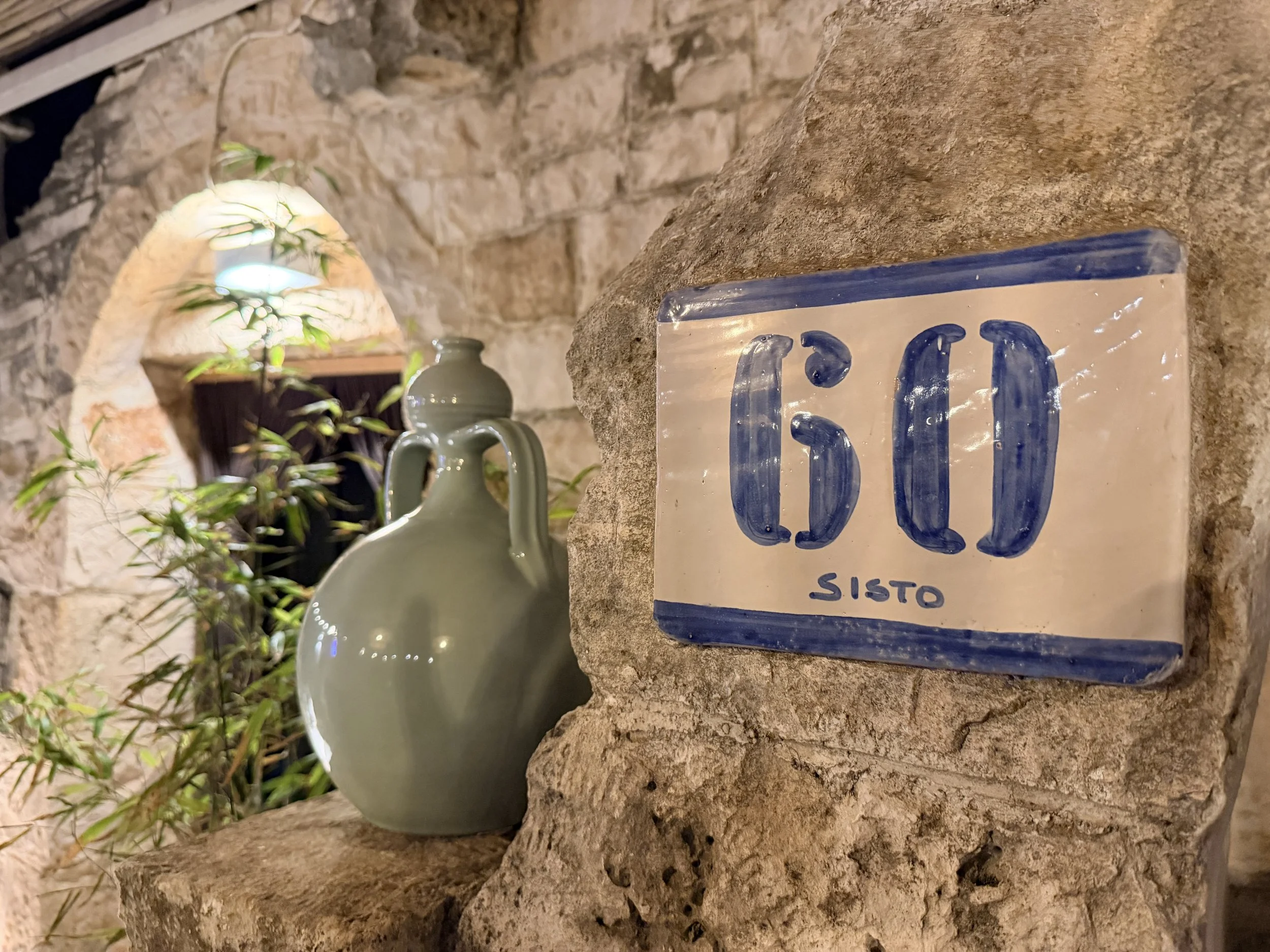 Piastrella con scritta '60 Sisto' attaccata su un muro di pietra, accanto a due vasi di ceramica verde chiaro, in un ambiente con pietre a vista.