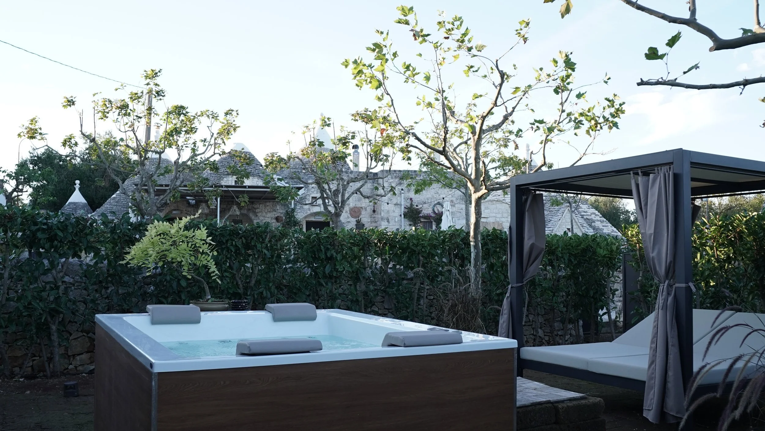 Piscina idromassaggio con cuscini e gazebo in giardino, con alberi e case di pietra sullo sfondo.